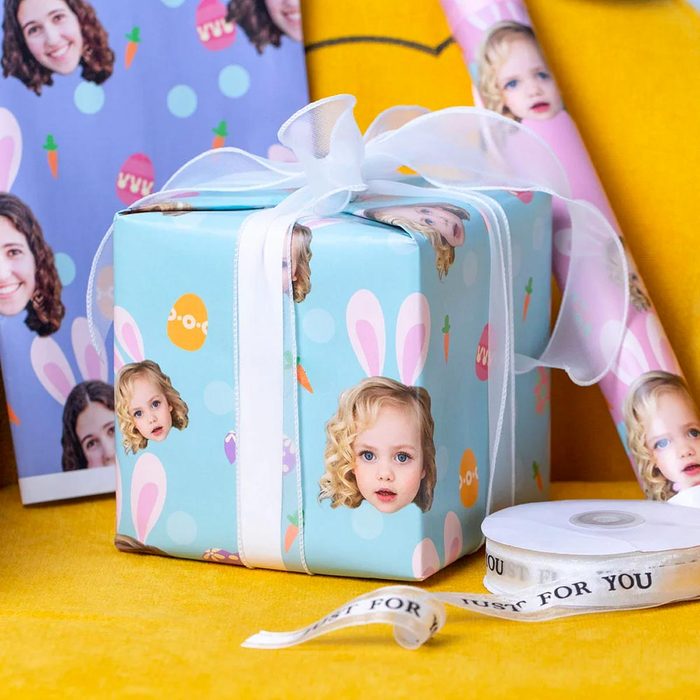 Gift box wrapped with personalized paper showing child faces, adorned with white ribbon; surrounded by rolls of similar wrapping paper.
