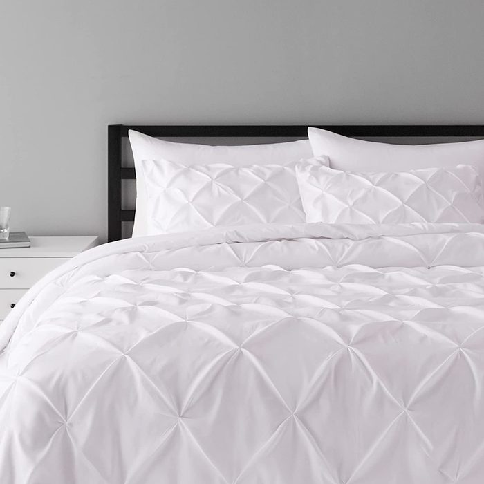 Bedding lies neatly on a bed, featuring a white, textured pattern, in a minimalist bedroom with a nearby nightstand holding a glass and book.