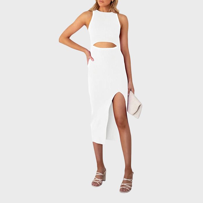 A woman stands, wearing a sleeveless white dress with a cutout and a slit, holding a textured clutch, on a plain background.
