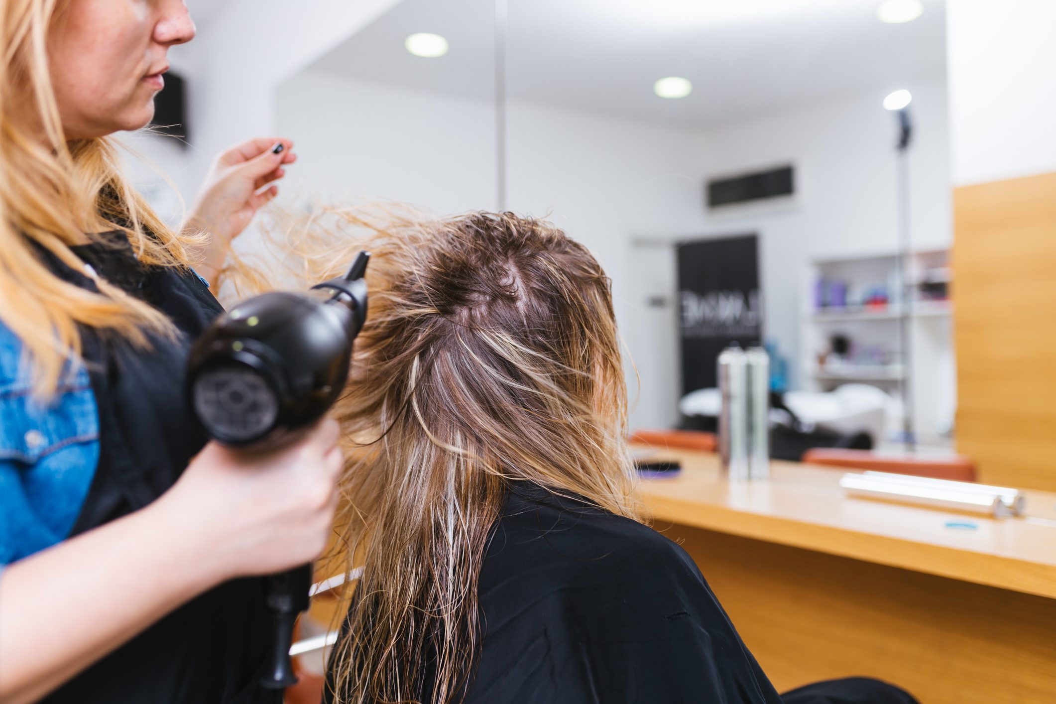 Person blow-dries hair; sits in front of salon mirror; styled indoor setting with beauty products visible on counter.
