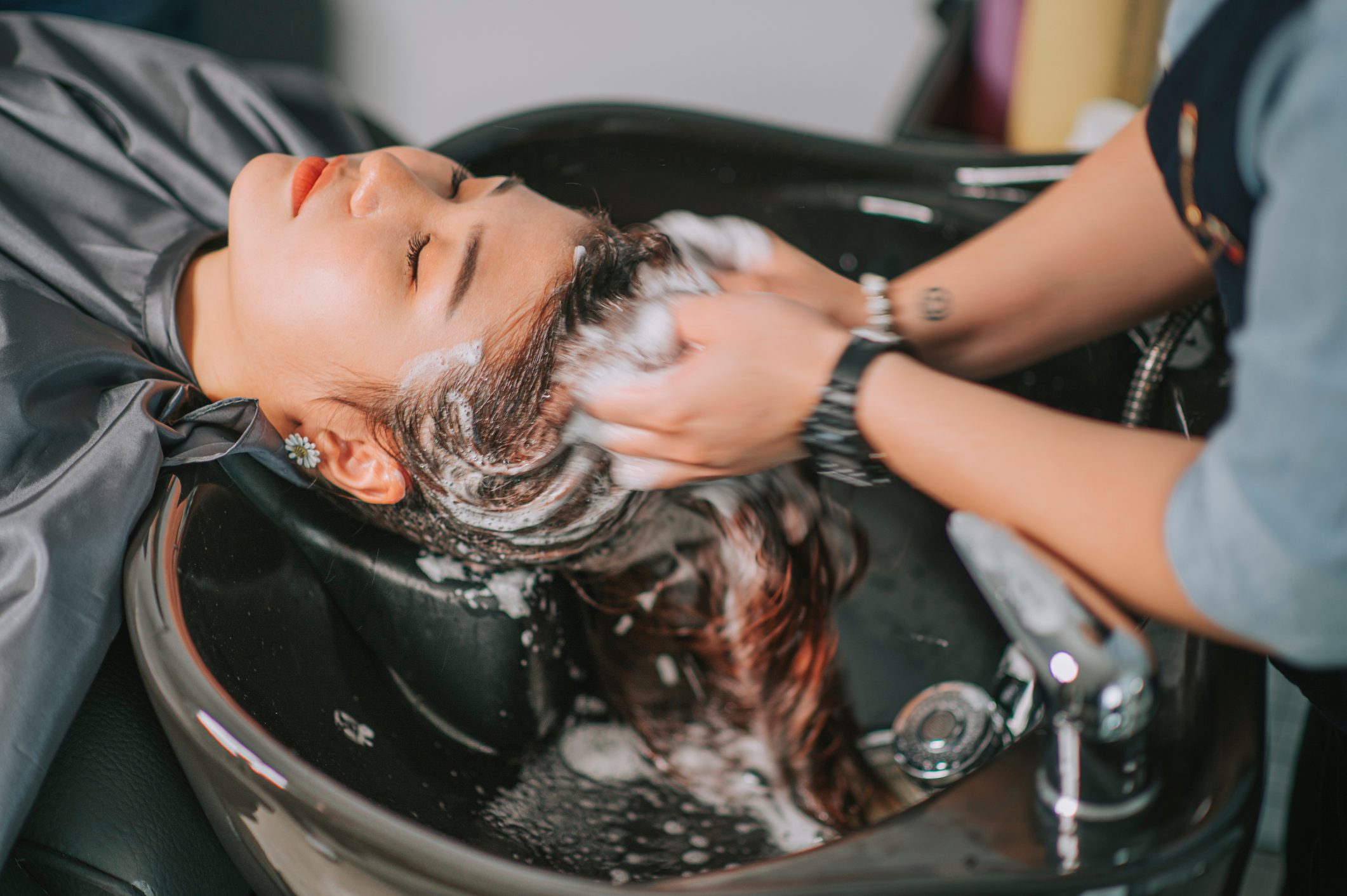 Person relaxes while receiving a hair wash in a salon setting, with hands massaging scalp and foamy shampoo visible.