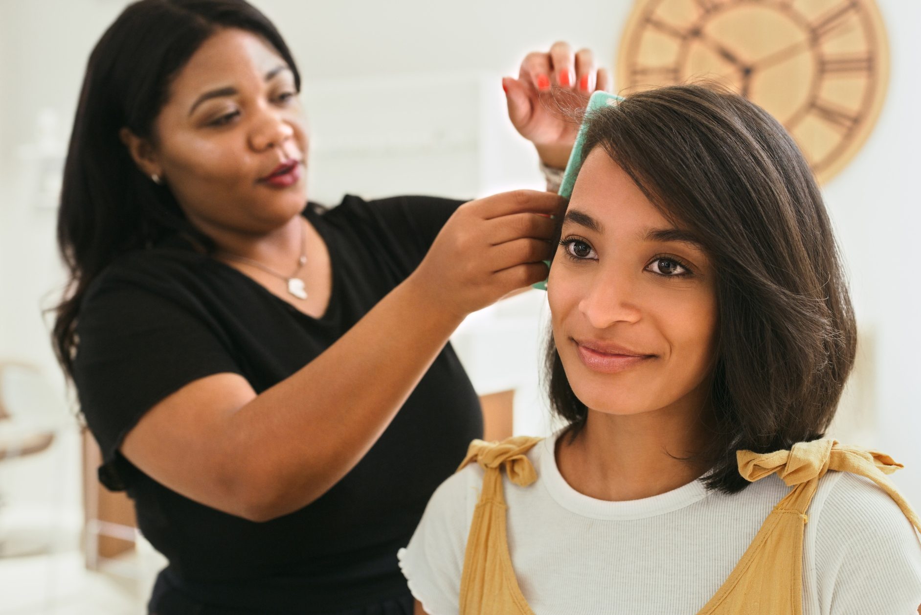 Stylist adjusts a woman's hair with a comb; both are in a salon with a large wall clock in the background.