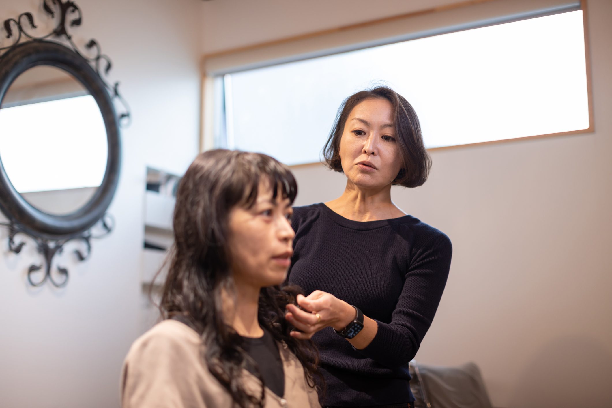 Stylist adjusting client's hair in a salon, decorated with a large ornate mirror and bright window light.
