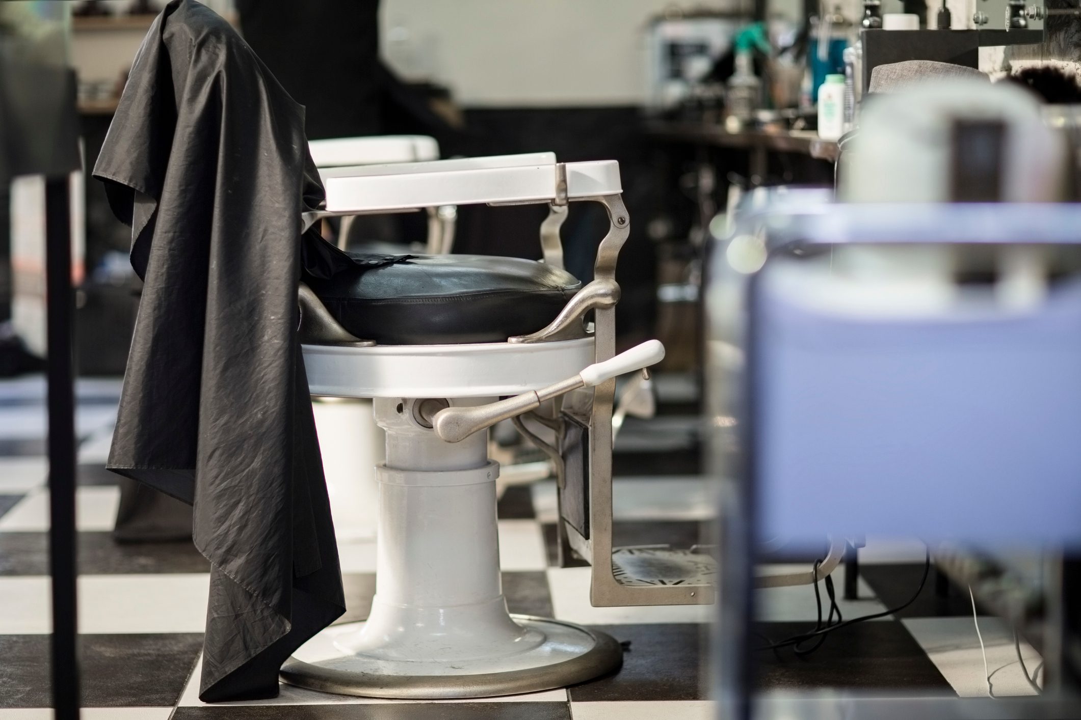 Barber chair holds draped cape, situated in a barbershop with black-and-white checkered floor, surrounded by hair products and equipment.