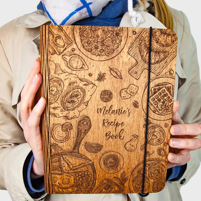 Wooden recipe book with engraved food illustrations held by a person in a coat. Text reads: 