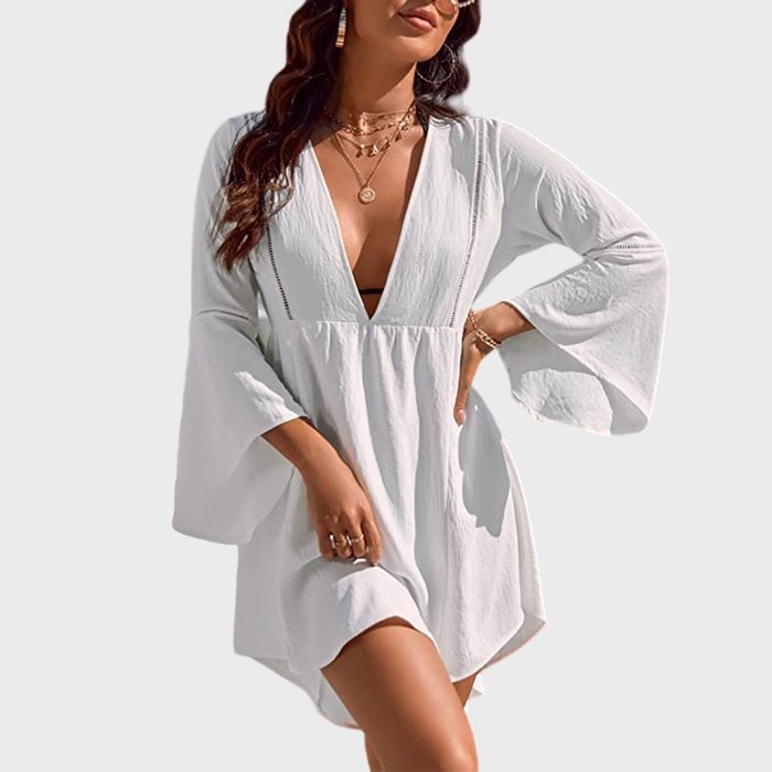 Woman wearing a white, deep V-neck dress, adorned with jewelry, posing against a plain background.