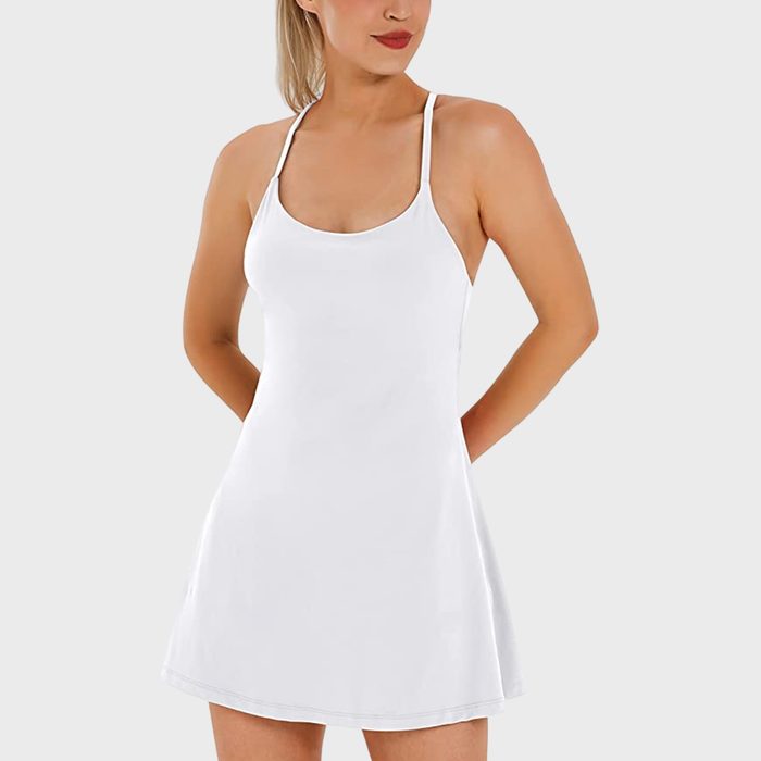 A woman stands wearing a white sleeveless dress, hands behind her back, posed against a plain light gray background.
