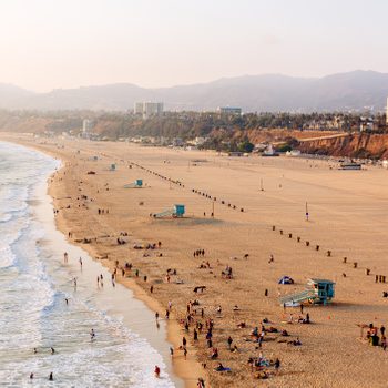 Beachgoers relax and swim along a sandy shoreline, with lifeguard towers and city buildings in the background under a hazy sky.