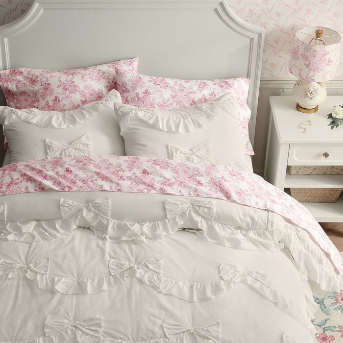 Bed displays ruffled white and pink floral linens; next to it, a white nightstand holds a floral lamp and small decorative items.