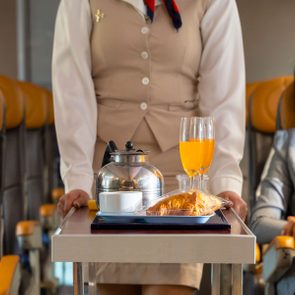 Tray holds kettle, orange juice, and croissant; attendant pushes it down narrow aisle in an airplane cabin with tan seats.