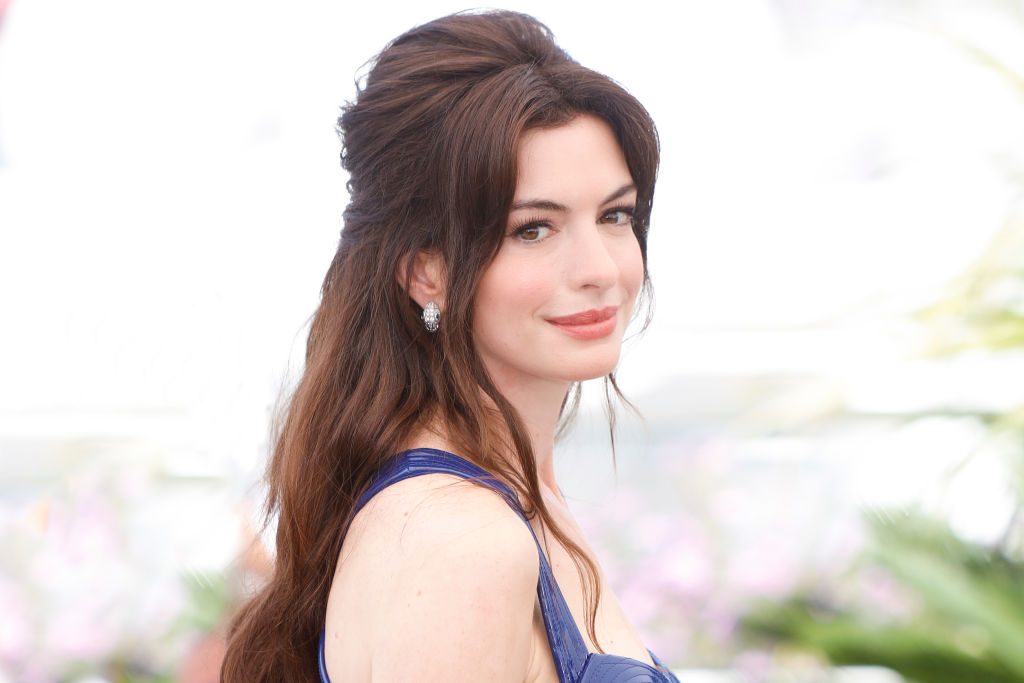 A woman with long dark hair smiles while wearing a blue dress, surrounded by a blurred, light-filled background with hints of greenery.