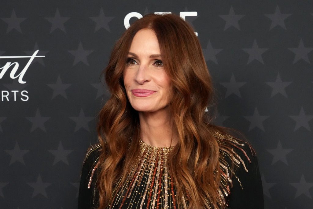 A woman with long, wavy hair smiles slightly, wearing a dress with gold embellishments, against a star-patterned backdrop.