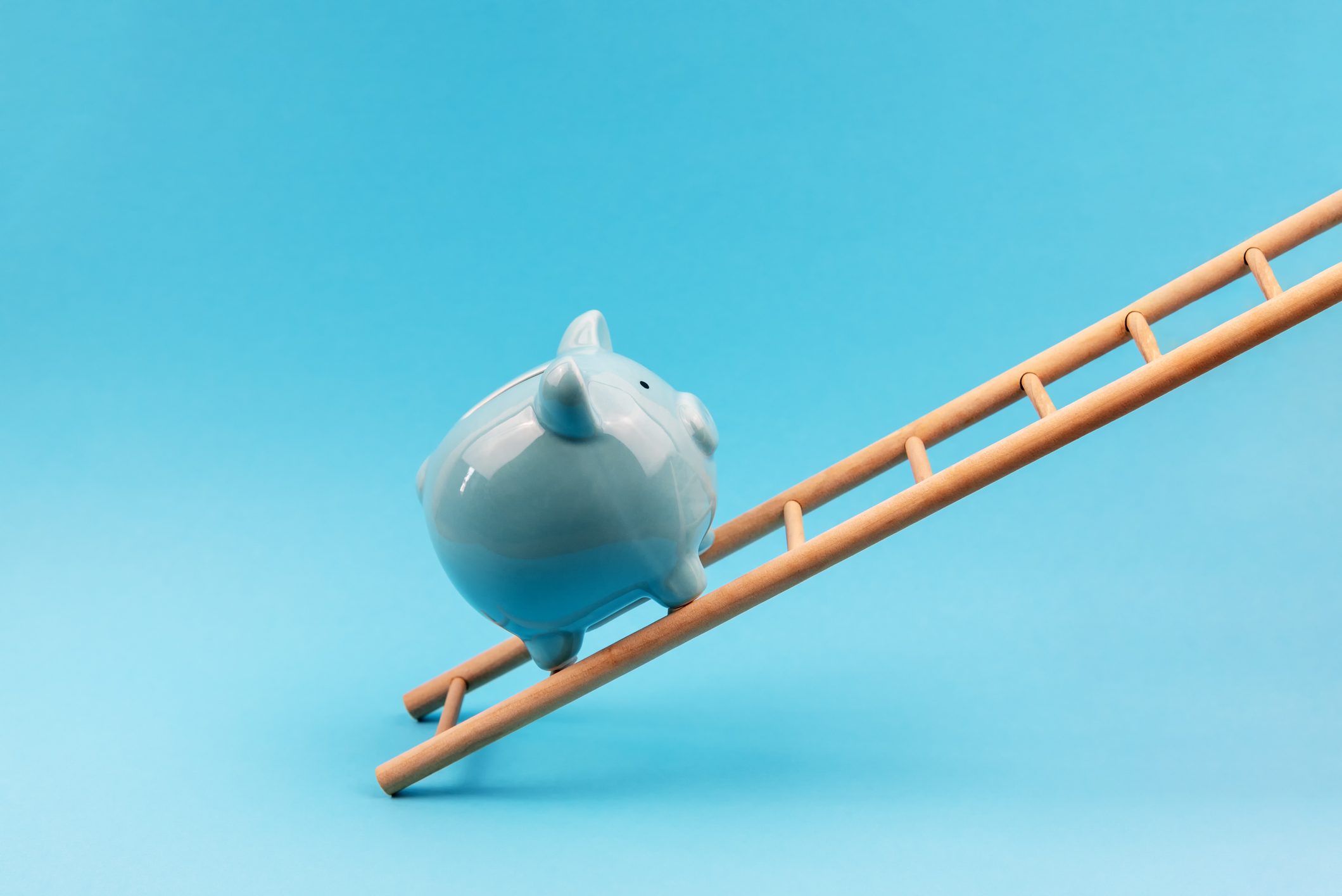 A blue piggy bank balances on an upward-slanting wooden ladder against a plain blue background.
