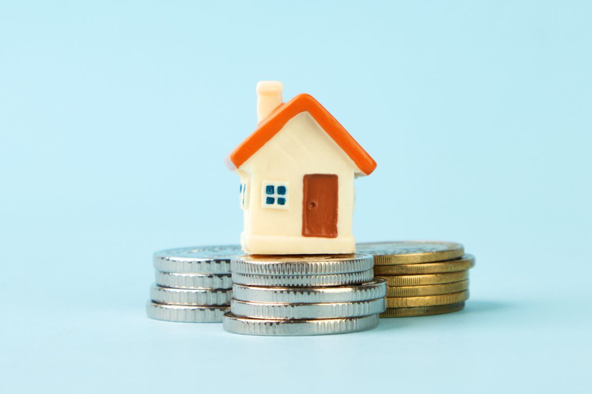 A small model house rests atop stacked coins, set against a plain blue background.