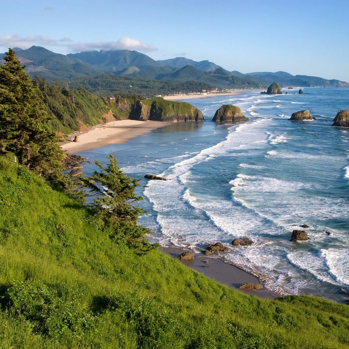 Waves crash on rocky shore beside a lush green cliff under a clear blue sky and distant mountains.