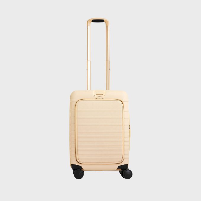 Suitcase stands upright, with telescopic handle extended, against a plain background. It has a beige color and four wheels for mobility.