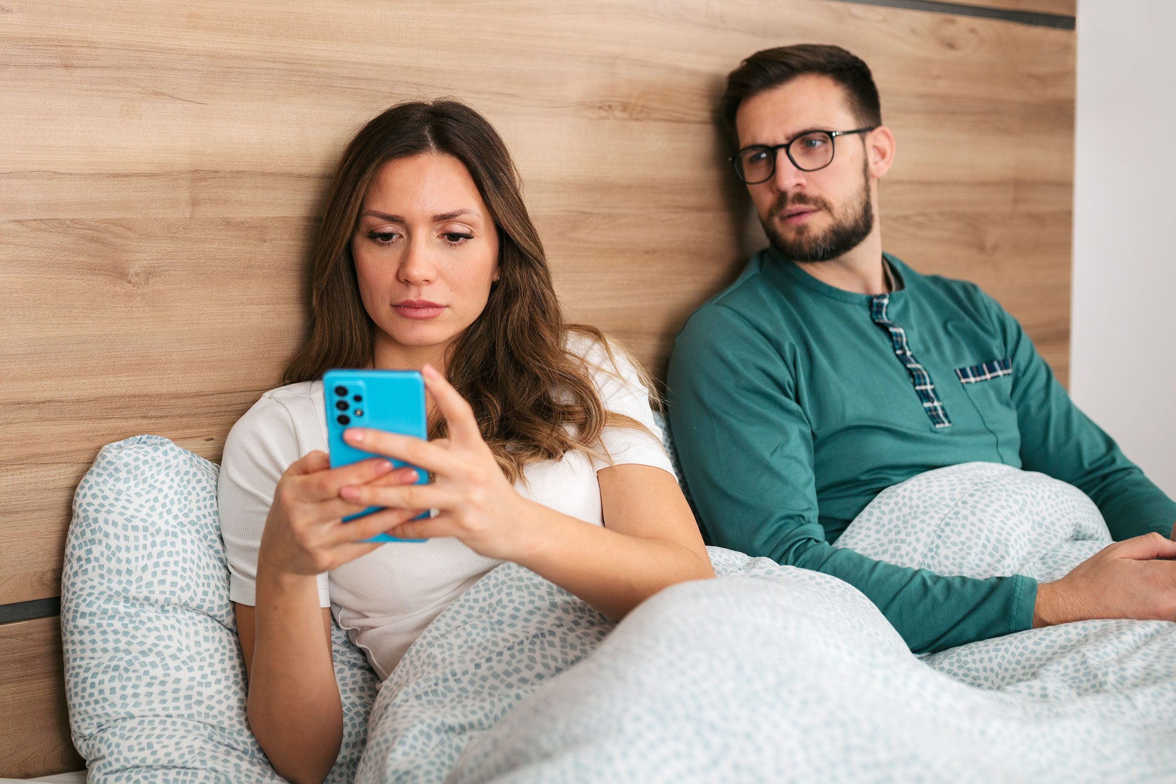 Woman using phone, sitting in bed. Man beside her looks over with questioning expression. Background features wooden headboard and patterned bedding.