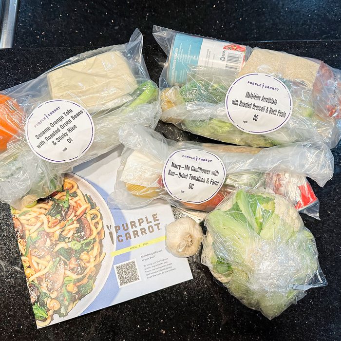Plastic bags contain labeled ingredients for three meals, accompanied by a Purple Carrot recipe card, all placed on a black countertop.