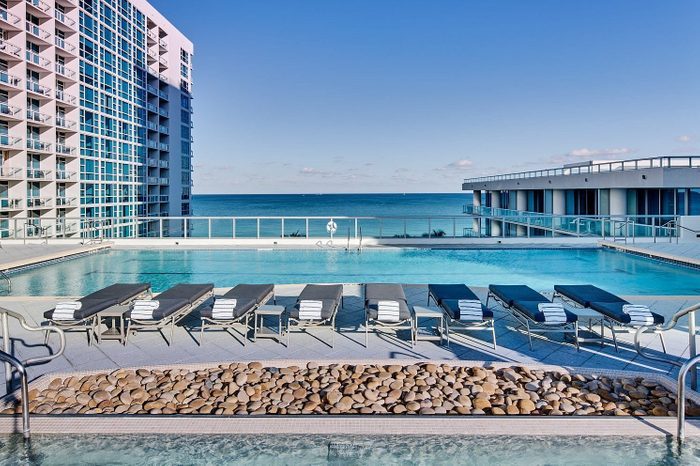 Poolside lounge chairs line up beside a pristine swimming pool overlooking the ocean, surrounded by modern high-rise buildings under a clear blue sky.