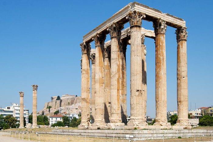 Ancient columns stand majestically amidst ruins, surrounded by a few scattered buildings and a clear blue sky in the background.