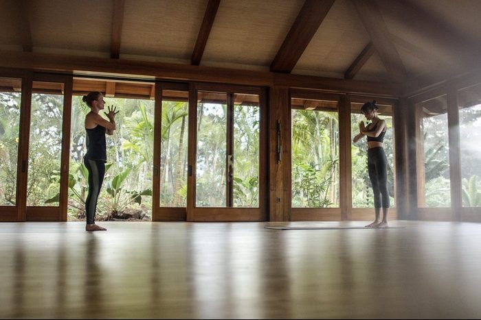 Two people stand in yoga poses inside a spacious room with large windows, surrounded by a lush garden outside.