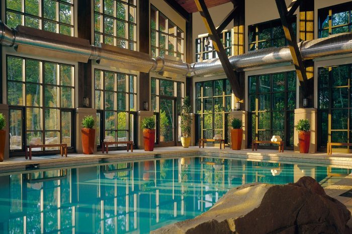 Indoor pool reflecting sunlight, surrounded by large windows and potted plants, with ductwork overhead and wooden benches along the perimeter.