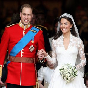 A couple walks arm-in-arm, smiling in formal wedding attire, surrounded by flower girls in a dimly lit venue.