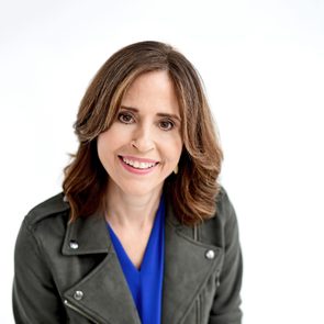 A person smiles at the camera, wearing a blue shirt and dark jacket against a plain white background.
