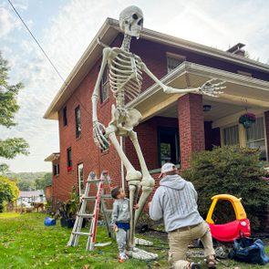 A tall skeleton stands while a child and adult adjust it beside a brick house, surrounded by a ladder and children's toys.