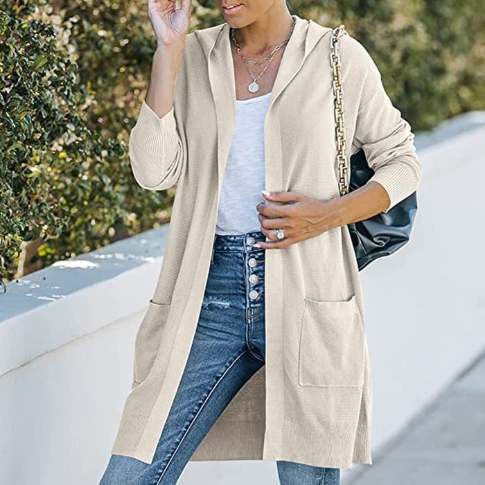 Person wearing a beige cardigan over a white shirt, holding a black bag, standing beside a white wall with greenery in the background.
