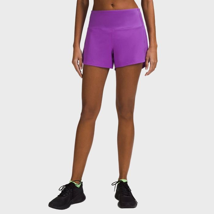 A person stands wearing purple shorts and black sneakers against a plain gray background.