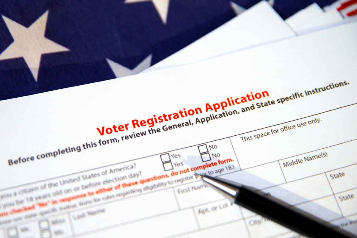 Form labeled "Voter Registration Application" resting on a table next to a pen, atop an American flag background.