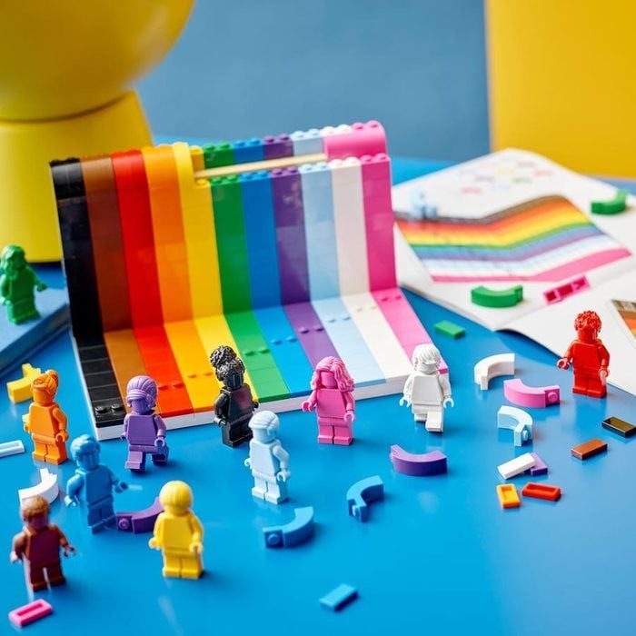 Lego figures stand before a rainbow-colored arch, surrounded by scattered bricks on a blue surface with an open instruction booklet nearby.