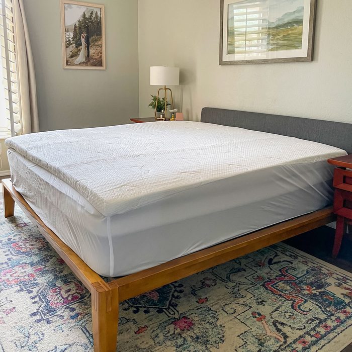 Bed with white mattress topper, positioned neatly in a bedroom. Wooden frame and nightstand; decorated with lamp, plant, art on gray walls.