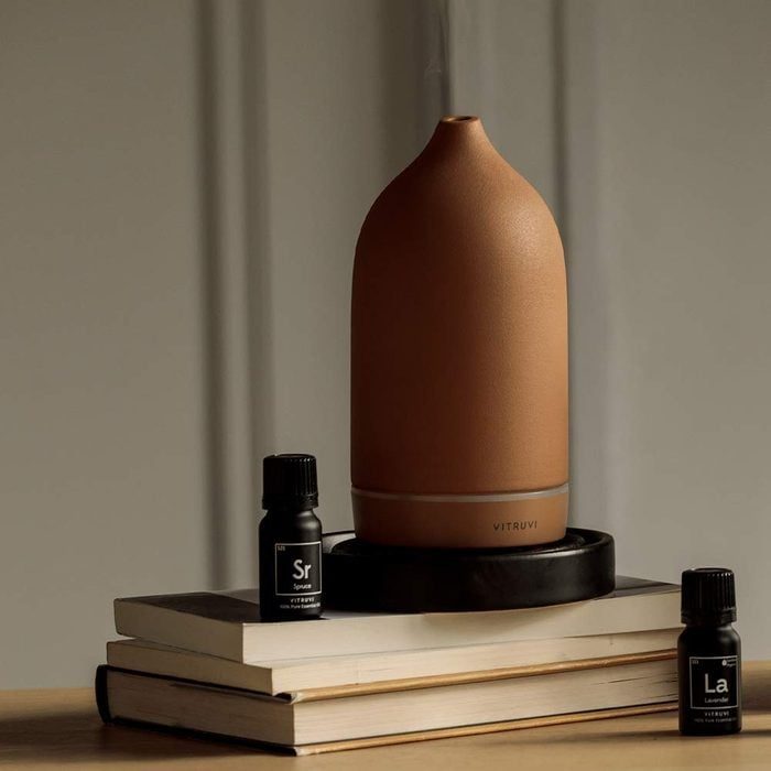 Diffuser releasing mist atop books beside essential oil bottles; background is a plain wall.
