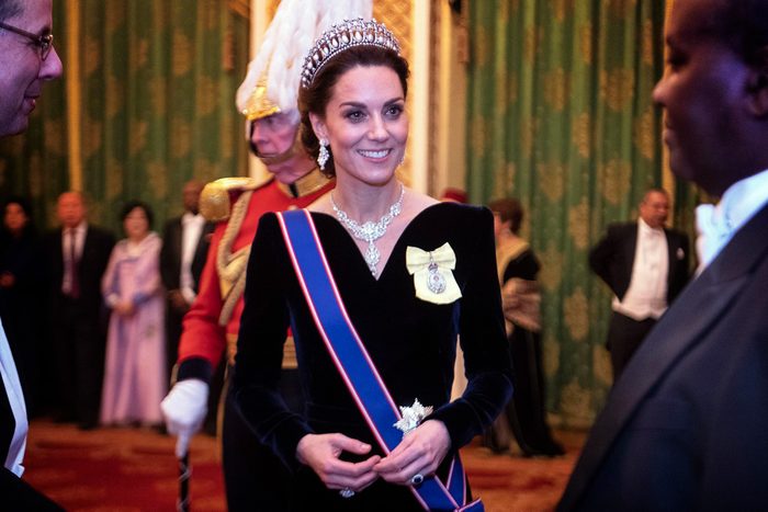 Kate Middleton wearing a tiara smiles in formal attire, engaging with people at a grand indoor event with elaborately decorated walls.