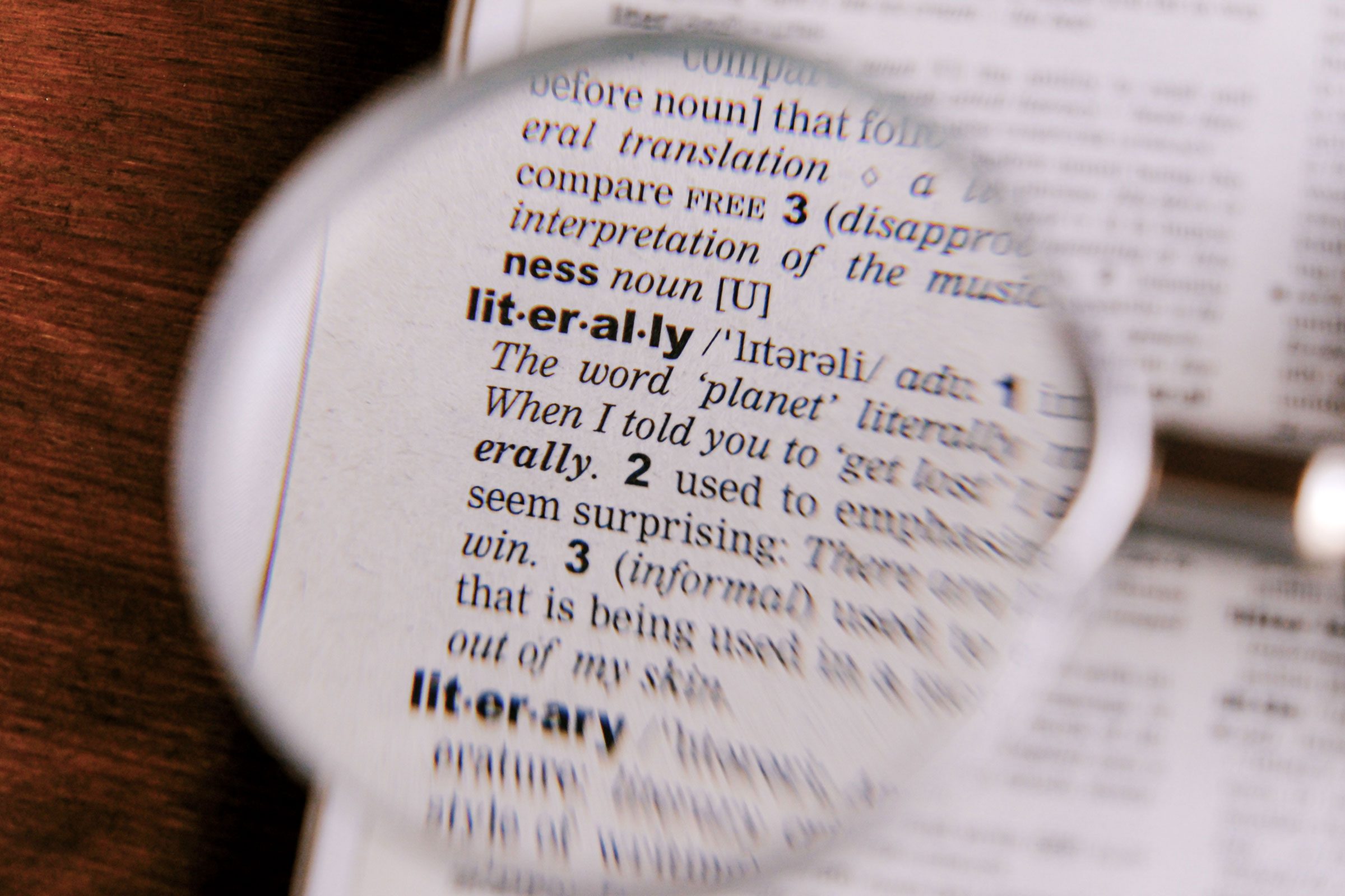 A magnifying glass placed over the word "literally" in a dictionary on a wooden surface.
