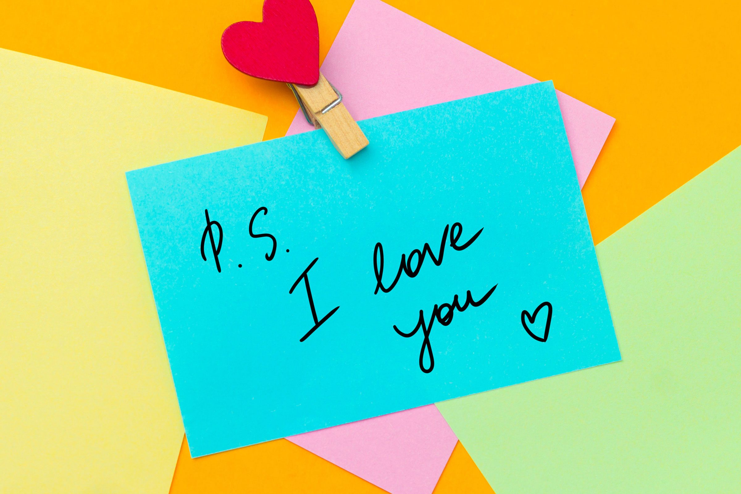 A lot of colored paper spread with a blue paper on top on which the text goes - "P.S. I LOVE YOU" with a heart peg.